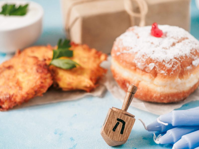Jewish holiday Hanukkah concept and background. Hanukkah food doughnuts and potatoes pancakes latkes, giftbox, candle and traditional spinnig dreidl on blue background. Copy space for text.